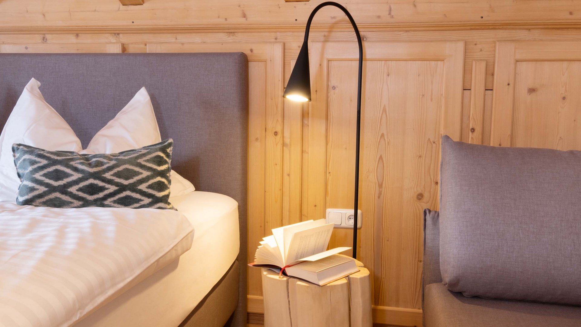 Hotel und Ferienwohnung im Allgäu, Oberstdorf Gemütliches Schlafzimmer mit Bett, Kissen, Nachttisch aus Baumstamm und Leselampe