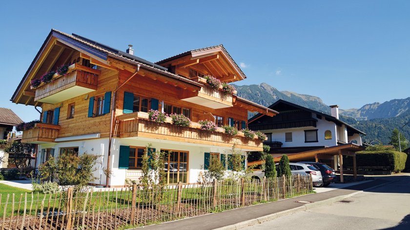 Hotel und Ferienwohnung im Allgäu, Oberstdorf Modernes Holzhaus mit Balkonblumen in Alpenlandschaft bei Sonnenschein