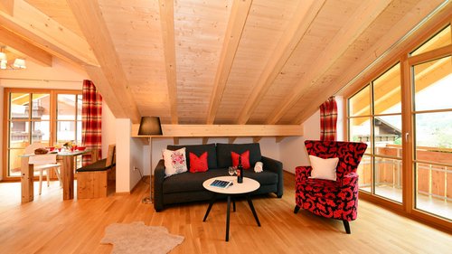 Wählt hier eure Ferienwohnung im Allgäu! Gemütliches Wohnzimmer mit Holzdecke, Sofa, rotem Sessel und Esstisch mit Fenster